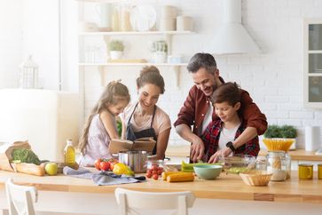 Glückliche Familie beim gemeinsamen Kochen in der Küche. Mutter und Tochter lesen Vater und Sohn das Rezept vor. Vater und Junge hacken grünes Gemüseblatt für Salat. Erholung und Lebensmittelzubereitung am Wochenende.