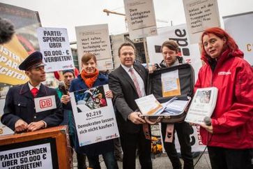 LobbyControl und Campact übergeben Bundesinnenminister Hans-Peter Friedrich (CSU) rund 100.000 Unterschriften für ein Verbot von Parteispenden über 50.000 Euro (pro Spender und Jahr) und für klare Regeln für Lobbyisten. Ein "Bahnmitarbeiter" trägt Friedrich symbolisch Gepäck hinterher, dass dieser im Zug zur großen Koalition liegen gelassen habe. Bild: "obs/Campact e.V./Jakob Huber"