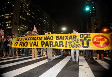 Demonstranten auf einer Straße von Rio de Janeiro. Auf dem Banner steht „Se a passagem não baixar, o Rio vai parar!“, übersetzt: „Wenn die Fahrkarten (-preise) nicht runtergehen, kommt Rio zum Stehen!“