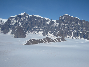 Die Watkins-Berge im südlichen Ostgrönland mit dem höchsten Gipfel Grönlands, Gunbjørn Fjeld (3,7 km über NN) im Hintergrund. Blick von ca. 2 km über Meereshöhe zum 1,5 km hohen Watkins-Steilabbruch, der sich in Basalte einschneidet, die am Übergang vom Paleozän zum Eozän vor 56 Mio. Jahren ausbrachen, damals nahezu auf Meereshöhe.