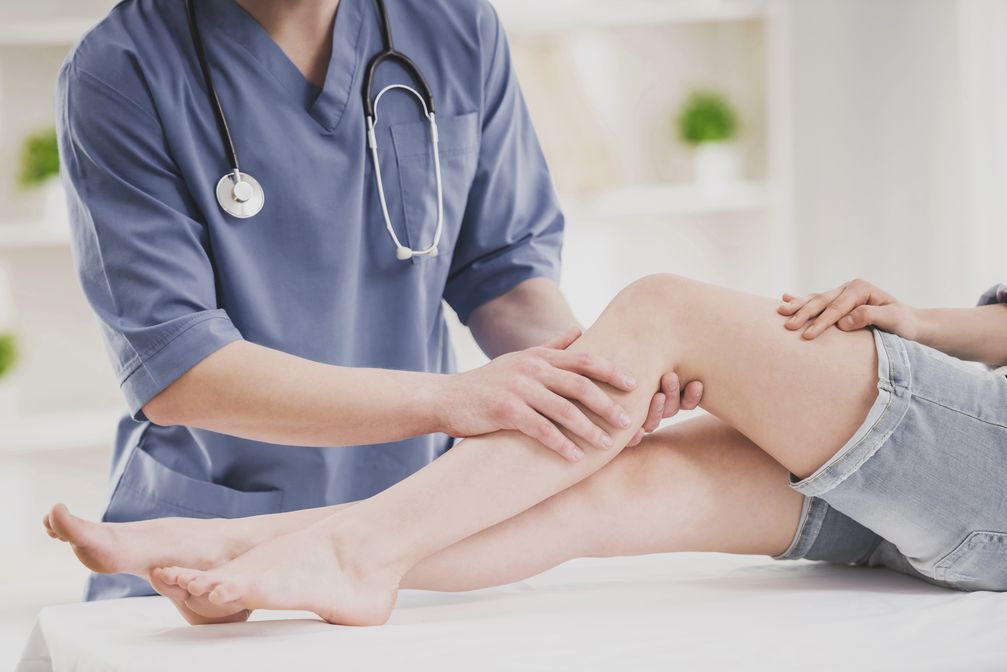 Close up. Doctor in Blue Uniform Comfforting Leg of Sitting Young Woman in Doctor Office. Traumatology Medical Devices. Medical Treatment in Clinic and Healthcare Professional Concept.