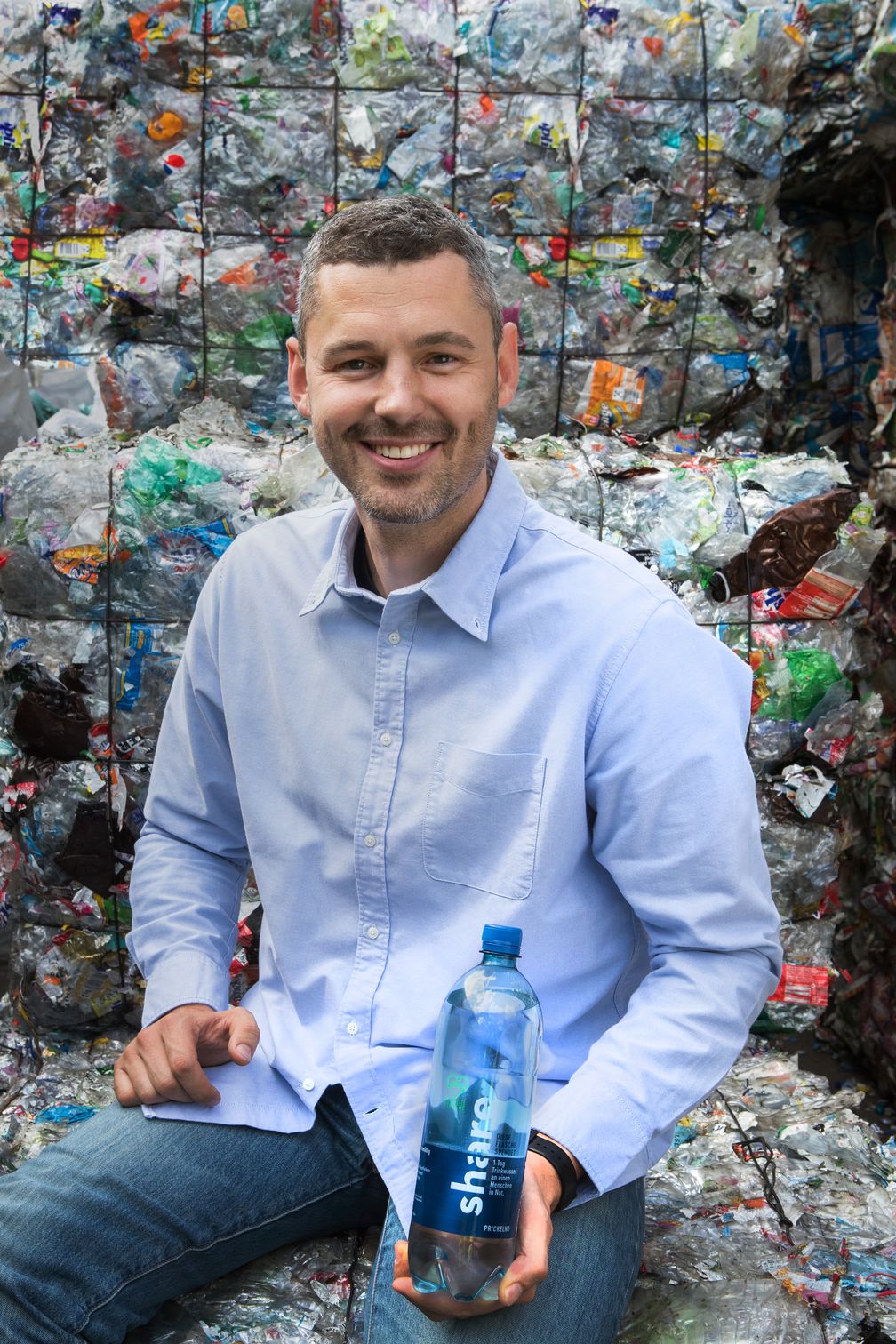 Dr. Sebastian Stricker mit der neuen "share"-Recyclatflasche vor dem Grundstoff seiner Innovation: Plastikmüll. Bild: "obs/REWE Markt GmbH/share"